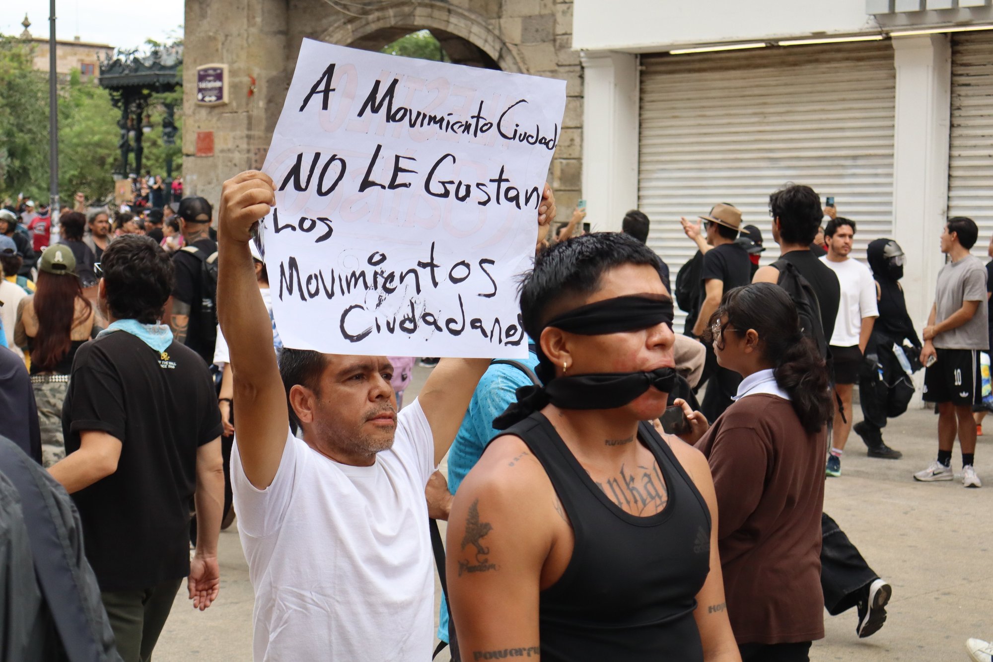 Un hombre levanta una pancarta que dice "A Movimiento Ciudadano no le gustan los movimientos ciudadanos" junto a un manifestante con ojos y boca cubiertos con tela negra en el Centro de Guadalajara